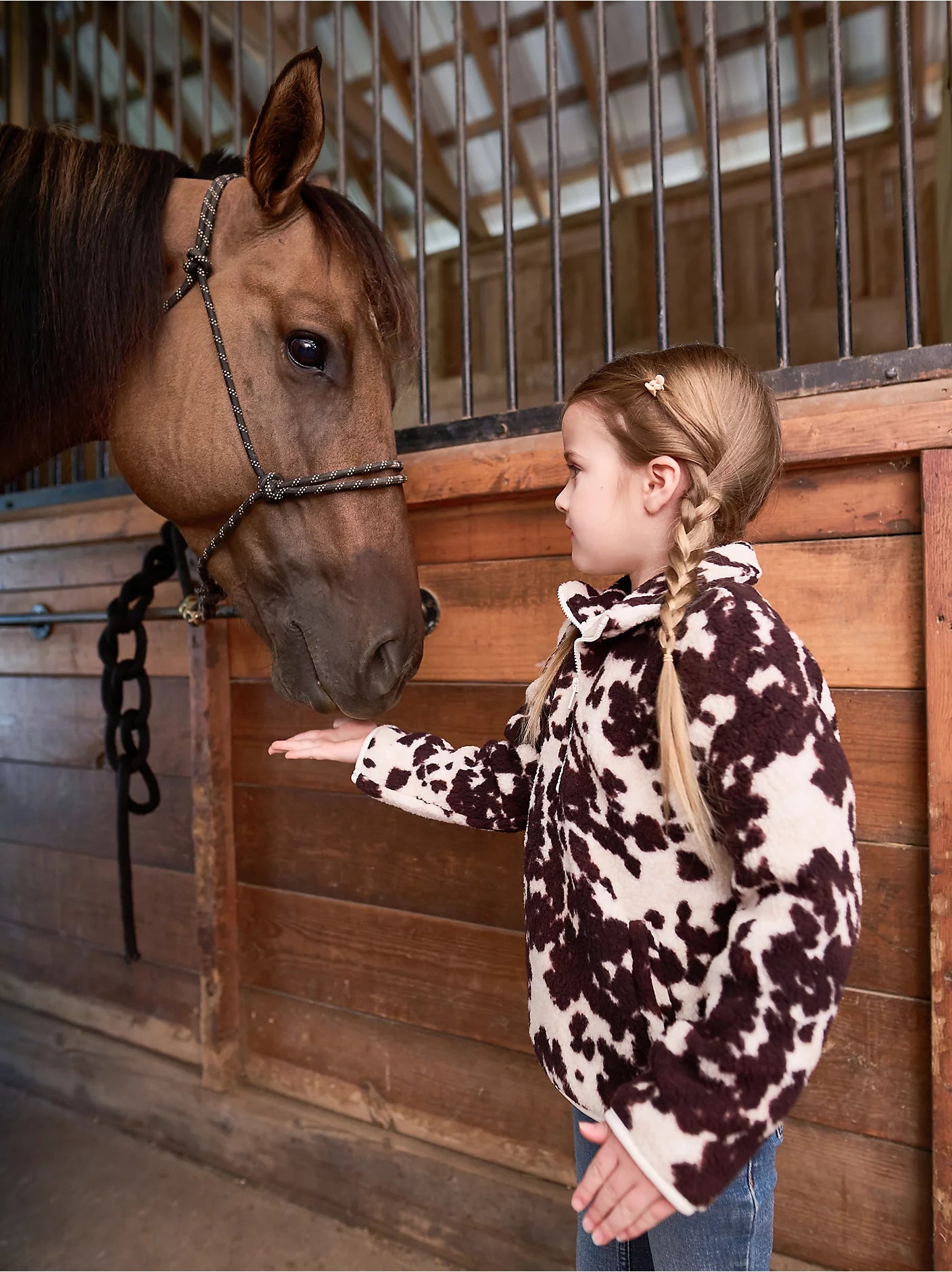 Sherpa vest for kids featuring a cozy cow print and a quarter zip, lifestyle shot in a barn with a horse.